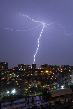 Dramatic vertical photograph capturing a powerful direct lightning strike hitting a residential building in night city. Branching electric discharge across the purple night sky above illuminated city