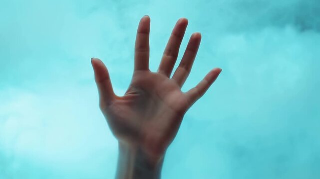 Blurred human hand silhouette behind blue glass with raised fingers in soft light