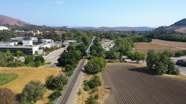 Aerial view of a passenger train travelling along tracks in a valley with suburban surroundings.