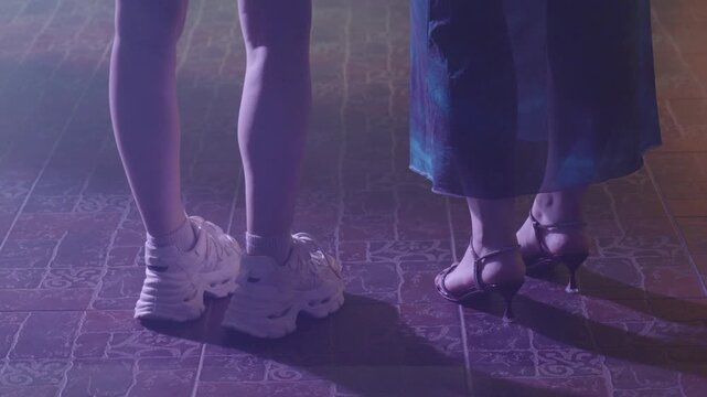 The lower legs of two women, one in chunky white sneakers and the other in high heels, stand on a tiled dance floor illuminated by colorful flashing lights.