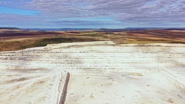The scene shows a large limestone quarry with different layers visible. Machinery is moving rock while workers are involved in operations under a clear sky during the day.