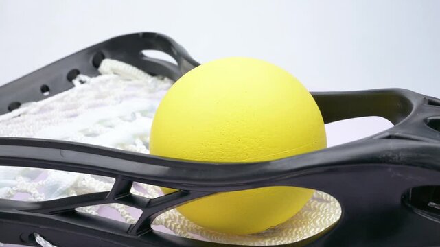 Bright yellow lacrosse ball resting inside a black lacrosse stick head with white woven netting rotating on white