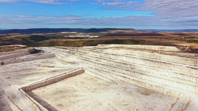 The scene shows a large quarry filled with earth. The ground is marked by dug-out shapes. In the background, hills rise against a clear blue sky. The area appears to be active in mineral extraction.