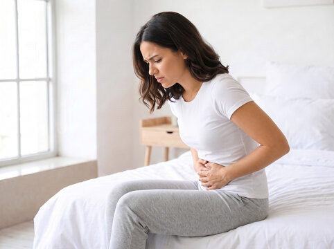 young hispanic woman sitting on bed holding stomach in severe abdominal pain caused by pmos menstrual cramps in home healthcare awareness concept