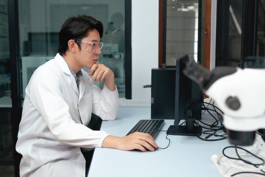 Young Asian scientist analyzing data on computer screen beside microscope in modern research laboratory