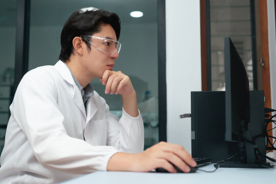 Young Asian scientist in lab coat analyzing data on computer screen thoughtfully in modern laboratory