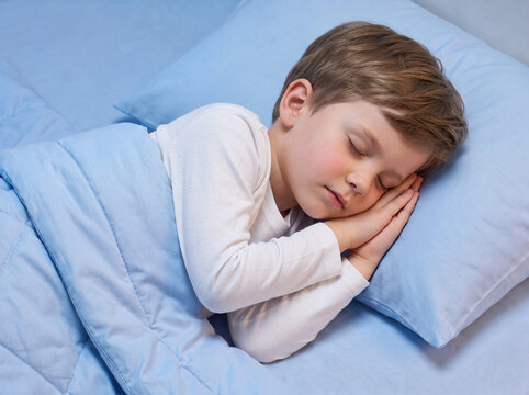 young caucasian boy peacefully sleeping on side in bed with hands under cheek on pastel sky blue cotton pillow and blanket in bedtime routine idea