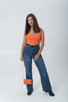 Young woman confidently modeling a casual and fashionable outfit, wearing an orange cropped top, blue jeans, and holding a small orange cross body bag on a clean white studio background