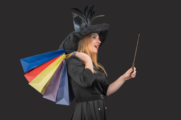 Woman dressed as a witch holding colorful shopping bags and a magic wand, looking excited while enjoying discounts and celebrating the Halloween holiday season against a dark background © somemeans