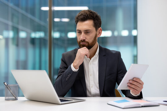 Serious young businessman in a suit sitting at an office desk, looking at a laptop and holding a tablet in his hand