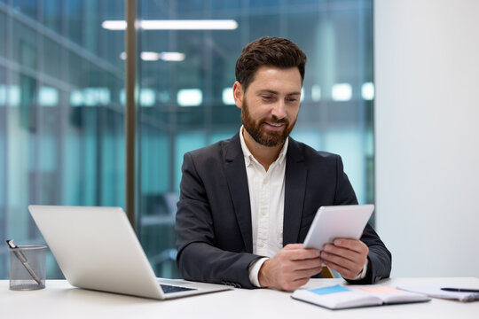 A young smiling businessman sitting at an office desk with a laptop, looking at the tablet he is holding in his hand
