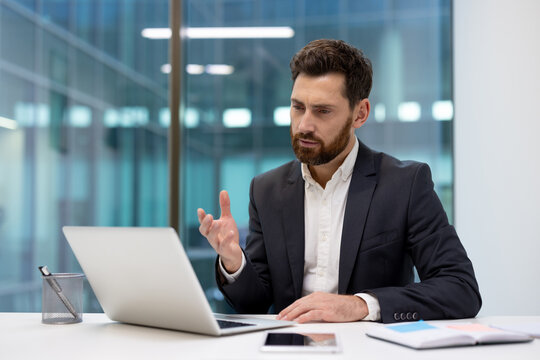 Serious young businessman sitting in the office at the table and talking on a video call on a laptop, smiling, gesturing with his hands