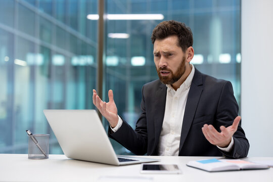 Angry young businessman in a suit sitting in the office, talking emotionally on a video call
