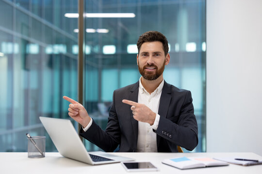 Portrait of a young businessman sitting in an office at a desk and pointing with his fingers at an empty space to the side