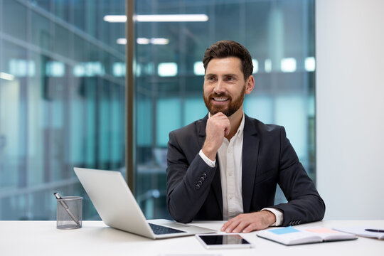Photo of a smiling young businessman in a suit sitting at a desk in the office, looking to the side