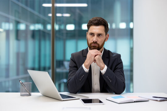 Photo of a serious young businessman in a suit sitting at a desk in the office, holding his hands together and looking to the side