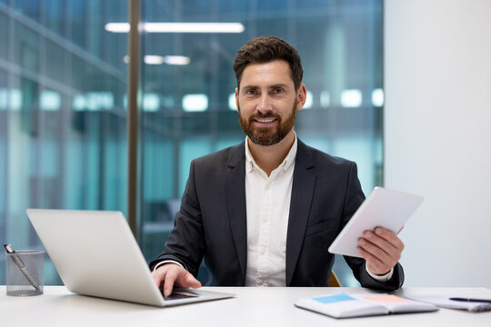 Portrait of a young smiling businessman sitting at an office desk with a laptop, holding a tablet in his hand and looking at the camera