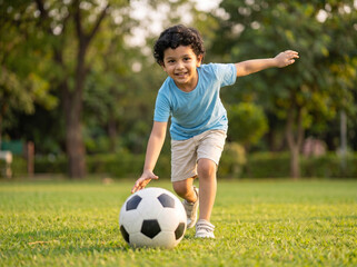 Fototapeta premium cheerful young indian boy playing with classic soccer football on lush green grass field in public park during warm evening golden hour sunlight