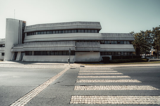 Weathered modernist building with long horizontal windows and angular concrete forms faces an empty road, where pale cobblestone crossing stripes lead toward the entrance and palm trees