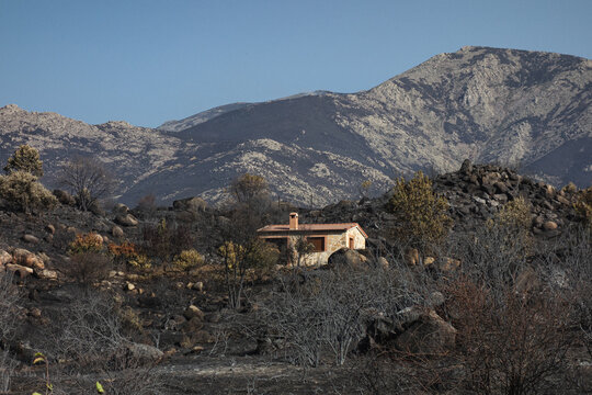 Resilient stone house standing amidst a charred landscape following a forest fire in Sierra Oeste de Madrid and Avila, Spain. Burnt vegetation and mountains under a clear sky