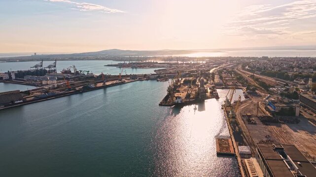 A port functions actively during sunset. Ships dock at the harbor while cranes operate. The city skyline and water reflect the orange light of the setting sun.