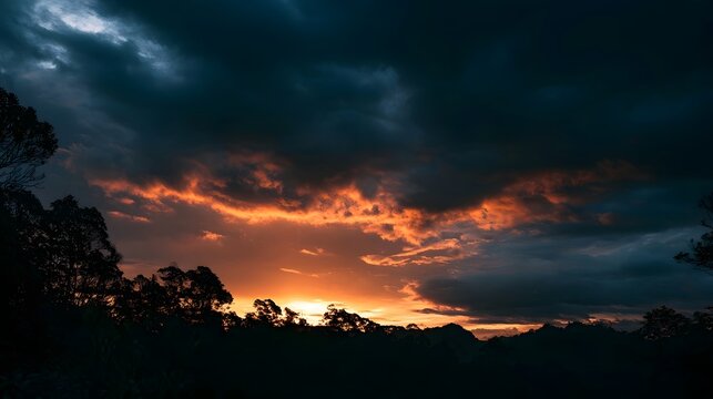 Dramatic sunrise with fiery orange clouds against dark stormy sky over silhouetted forest and hills