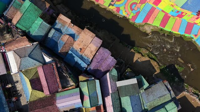 Top-down drone view over colorful Jodipan Village in Malang, Indonesia, showing painted rooftops, riverside murals, bridge and river in a vibrant urban neighborhood.