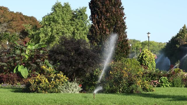 Fixed camera view of a traditional rotating sprinkler completing a full cycle while watering a landscaped garden, flowers and green lawn.
