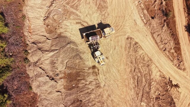 Two construction machines are at work on a dirt site, moving and loading soil. The sun is shining down on the area, which is mostly barren and dusty.