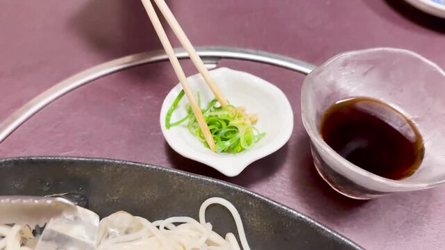 Chopsticks picking up green onions and putting them into soy sauce bowl for cold somen noodles meal.