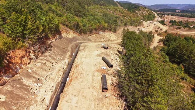 Workers are constructing a pipeline on a hillside surrounded by trees. The area shows earth being moved and sections of pipe lying on the ground. The time of day appears to be midday.