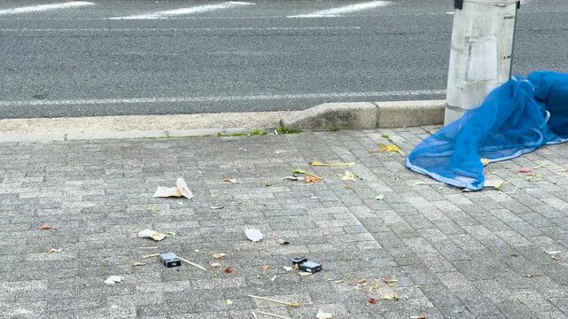 Person rides bicycle past littered sidewalk with blue tarp next to street.