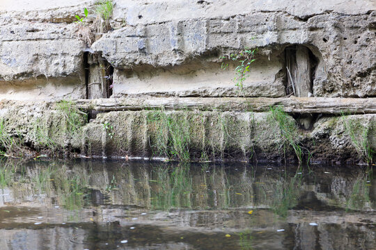 An old concrete dam with wooden elements and signs of decay above the water surface. Green plants grow through cracks in the concrete, while the quiet water reflects the texture of this old hydraulic 