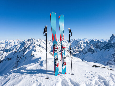 mountain skis with poles standing in deep snow on top of mountain with clear blue sky and snowy mountain range on background