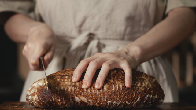 Close-up of baker in rustic linen apron slicing freshly baked artisan sourdough bread with knife on wooden cutting board. Homemade crusty loaf for breakfast, bakery promos, and healthy food concepts.