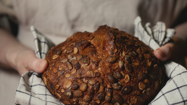 Close-up of baker in a rustic linen apron holding freshly baked artisan bread topped with pumpkin seeds on a checkered towel. Homemade loaf for bakery promos, organic product and healthy food concept.