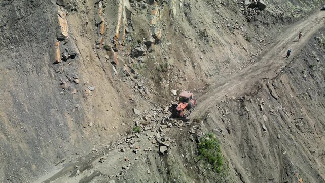 Aerial drone footage of an excavator working on a mountain road and removing rocks and loose debris from the base of a steep rocky slope.