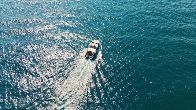 A small boat travels across the surface of the clear blue water during daytime. Sunlight reflects off the waves, creating shiny patterns in the water.