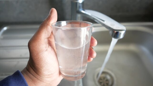Close up video of person holding glass of water and running tap in the background, showing clean water flowing into sink