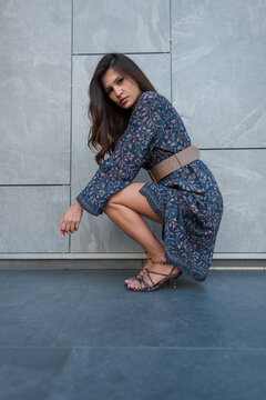 Side profile fashion portrait of a stylish young brunette woman crouching against a modern grey stone wall, wearing a floral boho dress, wide belt and high heels, looking at camera.
