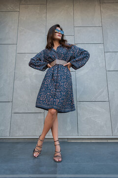 Stylish woman in a patterned blue midi dress, wide belt, sunglasses, and statement heels strikes a dynamic, low-angle pose against a modern grey tiled wall. Street fashion.