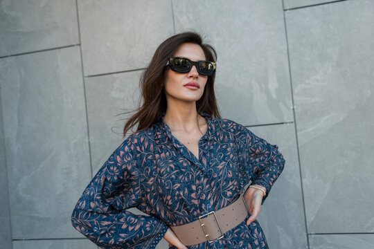A stylish young woman with sunglasses and a detailed floral print dress, belted at the waist, poses confidently with hands on hips against a modern grey tiled wall in daylight