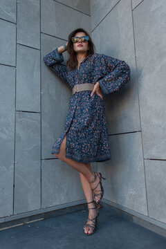 Confident woman in a blue patterned midi dress, wide tan belt, and leopard-print heels, poses against a modern grey building corner in an urban setting. Low-angle portrait.
