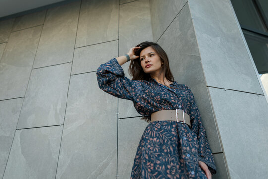 Low angle portrait of stylish brunette woman touching her hair near modern grey tile wall outdoors. Elegant female model wearing patterned dress in city street.