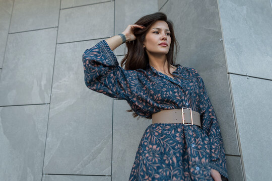 Urban trendy portrait of attractive brunette woman in patterned dress touching hair near modern grey wall. Stylish female fashion model posing outdoors in city.