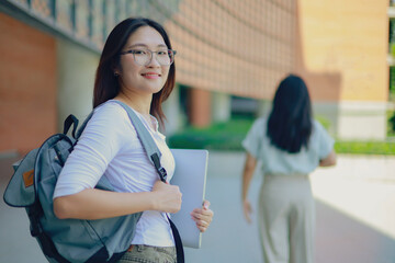 Happy inspiring Asian female undergraduate student with backpack and textbooks in her hands stands in front of a university building looking and smiling at the camera. © ฺฺฺBoonterm