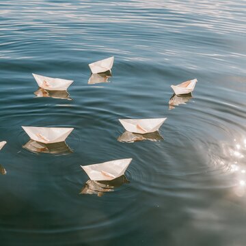 Paper boats floating on calm blue water, one drifting alone with a soft reflection for anti bullying and exclusion concepts