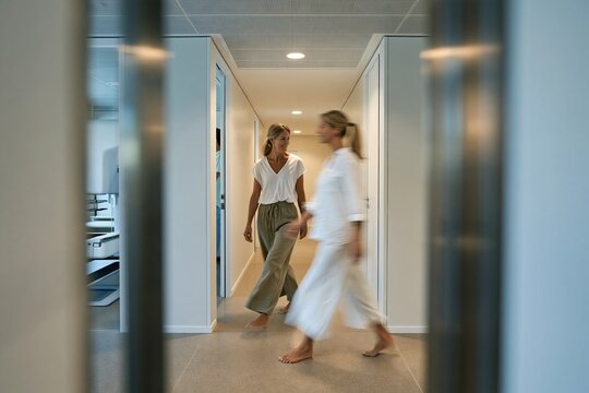 Two women walking through a soft, dusty rose lit mammogram screening corridor with a calm, airy atmosphere for healthcare and wellness
