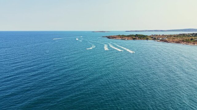 Several boats create wake patterns in the water near a coastal area with land visible in the background. The sky is clear, and it is a bright day.