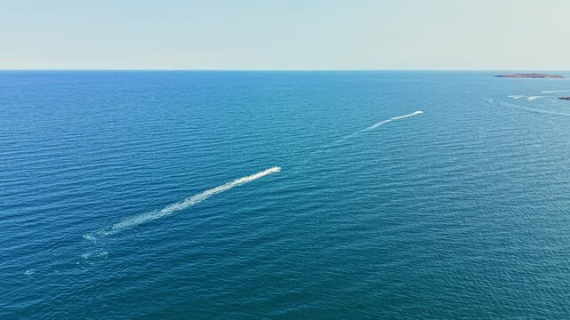 Two boats travel across the blue sea, leaving trails behind them. The sky is clear and the waves are visible on the water surface. It is daytime and calm.